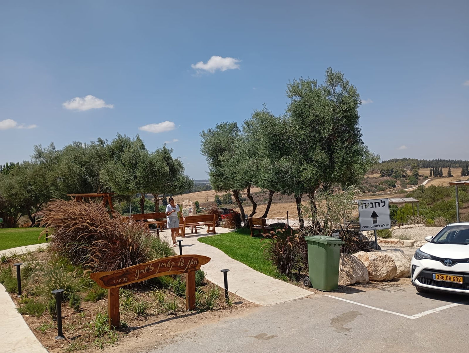 פינת ישיבה מוצלת בעצי זית, שביל גישה, שלט חניה וגינה ירוקה במשתלה ישראלית